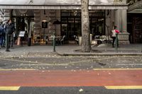pedestrians walk past a cafe in a downtown area with a large tree in the middle