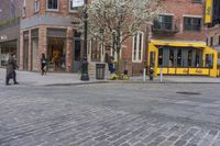 a red brick street in the middle of downtown area in a city with two people, and a yellow trolley in front