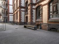 a bench sits in the middle of a courtyard with parked bicycles nearby and tall brick buildings