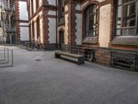 a bench sits in the middle of a courtyard with parked bicycles nearby and tall brick buildings