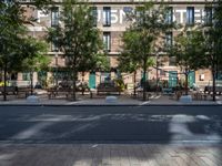 a bunch of tables on the sidewalk near buildings on the other side of it and trees behind