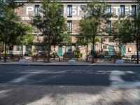 a bunch of tables on the sidewalk near buildings on the other side of it and trees behind