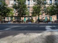 a bunch of tables on the sidewalk near buildings on the other side of it and trees behind