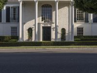 a large white mansion has black shutters and three doors and many bushes on the sidewalk