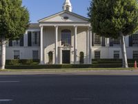 a large white mansion has black shutters and three doors and many bushes on the sidewalk