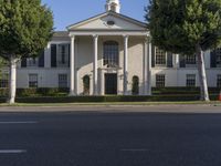 a large white mansion has black shutters and three doors and many bushes on the sidewalk