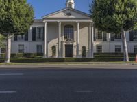 a large white mansion has black shutters and three doors and many bushes on the sidewalk