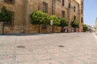 a man walks down a paved street past stone buildings and orange trees that sit on either side