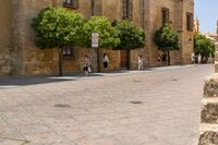 a man walks down a paved street past stone buildings and orange trees that sit on either side