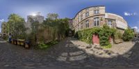 a fisheye lens view of buildings, bushes, and trash cans on a cobble - stone street