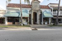 a street is lined with shops and palm trees on either side of it, and some are with large awnings