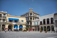 two men riding horses around a plaza while a lady stands nearby on the sidewalk at the end of the square