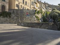 a street sign is in front of a stone wall by some trees and buildings and a stone walkway