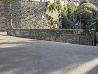 a street sign is in front of a stone wall by some trees and buildings and a stone walkway