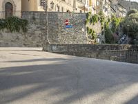 a street sign is in front of a stone wall by some trees and buildings and a stone walkway