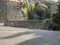 a street sign is in front of a stone wall by some trees and buildings and a stone walkway