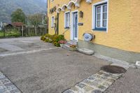 the front of the house has a yellow painted wall with white trim and windows and two flowers in blooming pots on the sidewalk