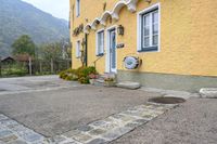 the front of the house has a yellow painted wall with white trim and windows and two flowers in blooming pots on the sidewalk