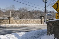 street signs on the corner next to a wall and road covered in snow, and trees