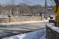 street signs on the corner next to a wall and road covered in snow, and trees