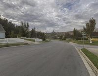 a street in front of a white fence and houses in the distance on a cloudy day