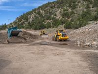 two yellow bulldozers work on sand and gravel in a mountain valley while another one looks at it