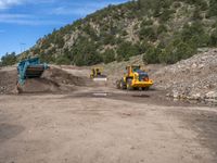 two yellow bulldozers work on sand and gravel in a mountain valley while another one looks at it