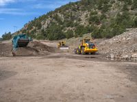 two yellow bulldozers work on sand and gravel in a mountain valley while another one looks at it