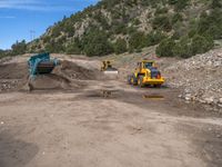 two yellow bulldozers work on sand and gravel in a mountain valley while another one looks at it