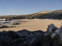 a man walking along a beach towards the water and sand of another beach with a large amount of rocks and mountains