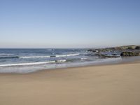 a large open beach and calm ocean waves crashing in front of a house and hill