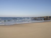 a large open beach and calm ocean waves crashing in front of a house and hill