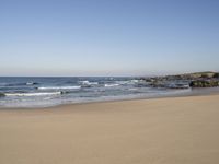 a large open beach and calm ocean waves crashing in front of a house and hill