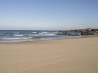 a large open beach and calm ocean waves crashing in front of a house and hill