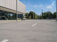 an empty street with parking meters and parked vehicles in front of a large building and sky