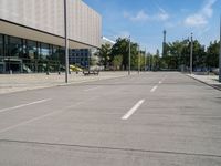 an empty street with parking meters and parked vehicles in front of a large building and sky