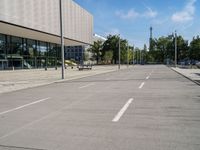 an empty street with parking meters and parked vehicles in front of a large building and sky