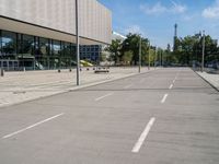 an empty street with parking meters and parked vehicles in front of a large building and sky