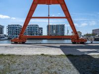 an orange crane on the docks looking out to the water with some buildings behind it