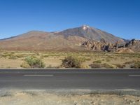 a road in front of a mountain and a dirt field with bushes and trees to the side