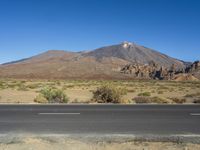 a road in front of a mountain and a dirt field with bushes and trees to the side