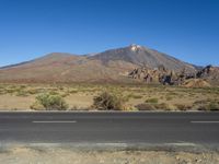 a road in front of a mountain and a dirt field with bushes and trees to the side