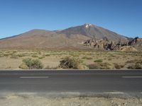 a road in front of a mountain and a dirt field with bushes and trees to the side