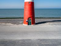 the lighthouse is bright red and white near the ocean water a black bench can be seen