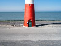 the lighthouse is bright red and white near the ocean water a black bench can be seen