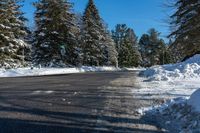 a road next to a pile of snow with pine trees in the background and a blue sky with clouds