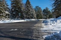 a road next to a pile of snow with pine trees in the background and a blue sky with clouds