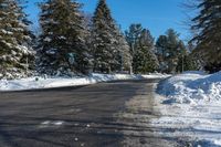 a road next to a pile of snow with pine trees in the background and a blue sky with clouds