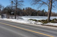 a road with snow and trees next to a street sign and a field of snow