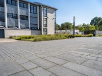 empty concrete parking lot with large buildings in back ground area of outdoor space next to tall grass and shrubs